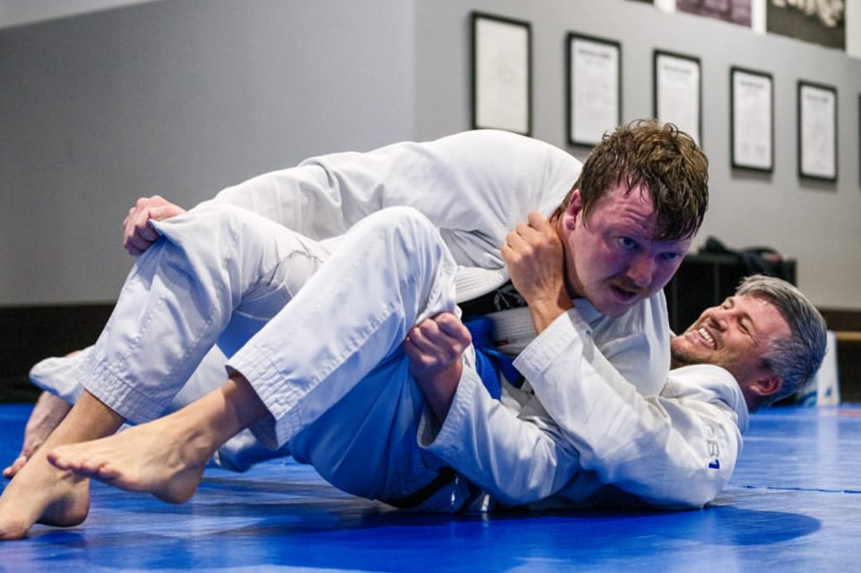 A Brazilian Jiu-Jitsu practitioner controls their opponent after passing guard during a sparring session, the bottom athlete is smiling.