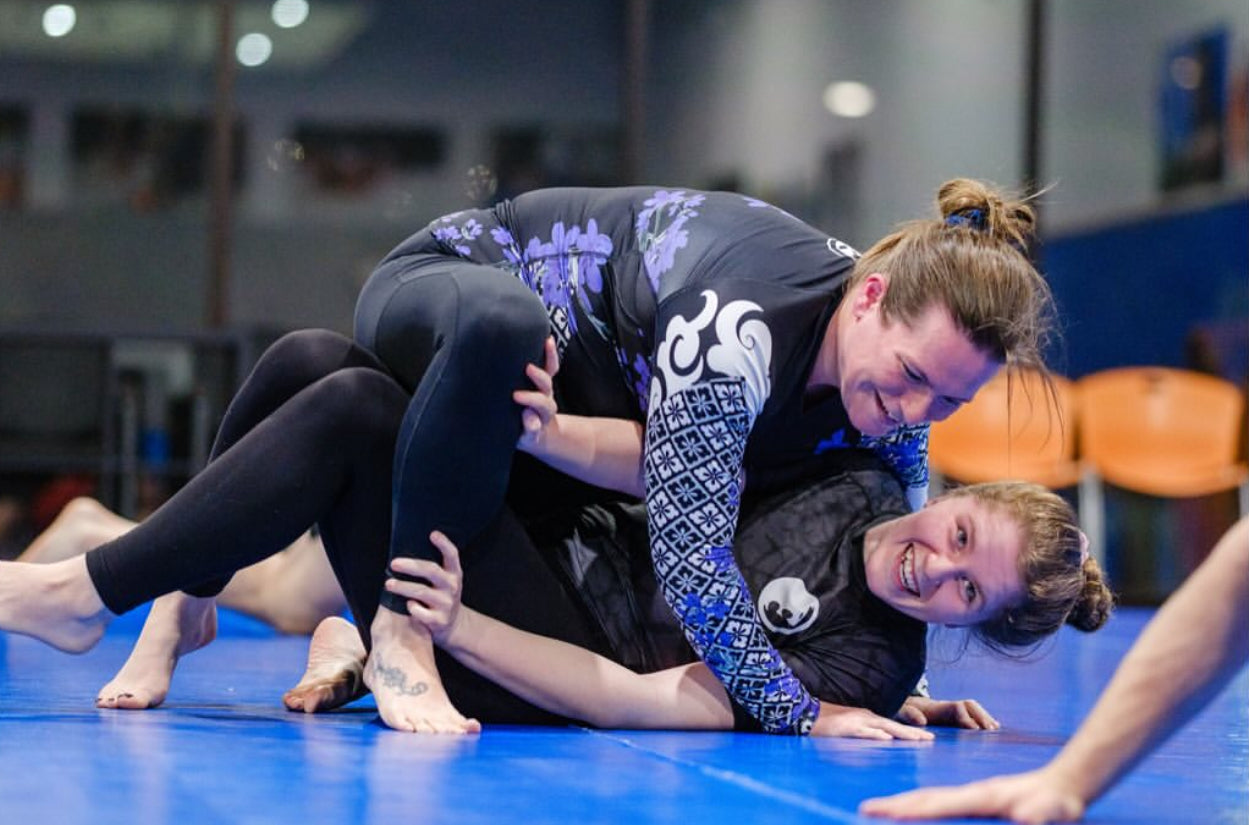 Two athletes roll on the mat in a high-energy jiu-jitsu sparring session, smiling as they battle for position.